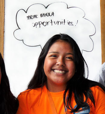 A photo of a girl smiling in an orange t-shirt with the words ask me about new opportunities written on a white board behind her