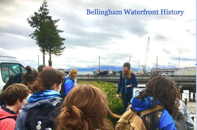 Instructor Anna Booker talks to a group of students standing near the Bellingham waterfront. On top of the image are the words Bellingham Waterfront History.