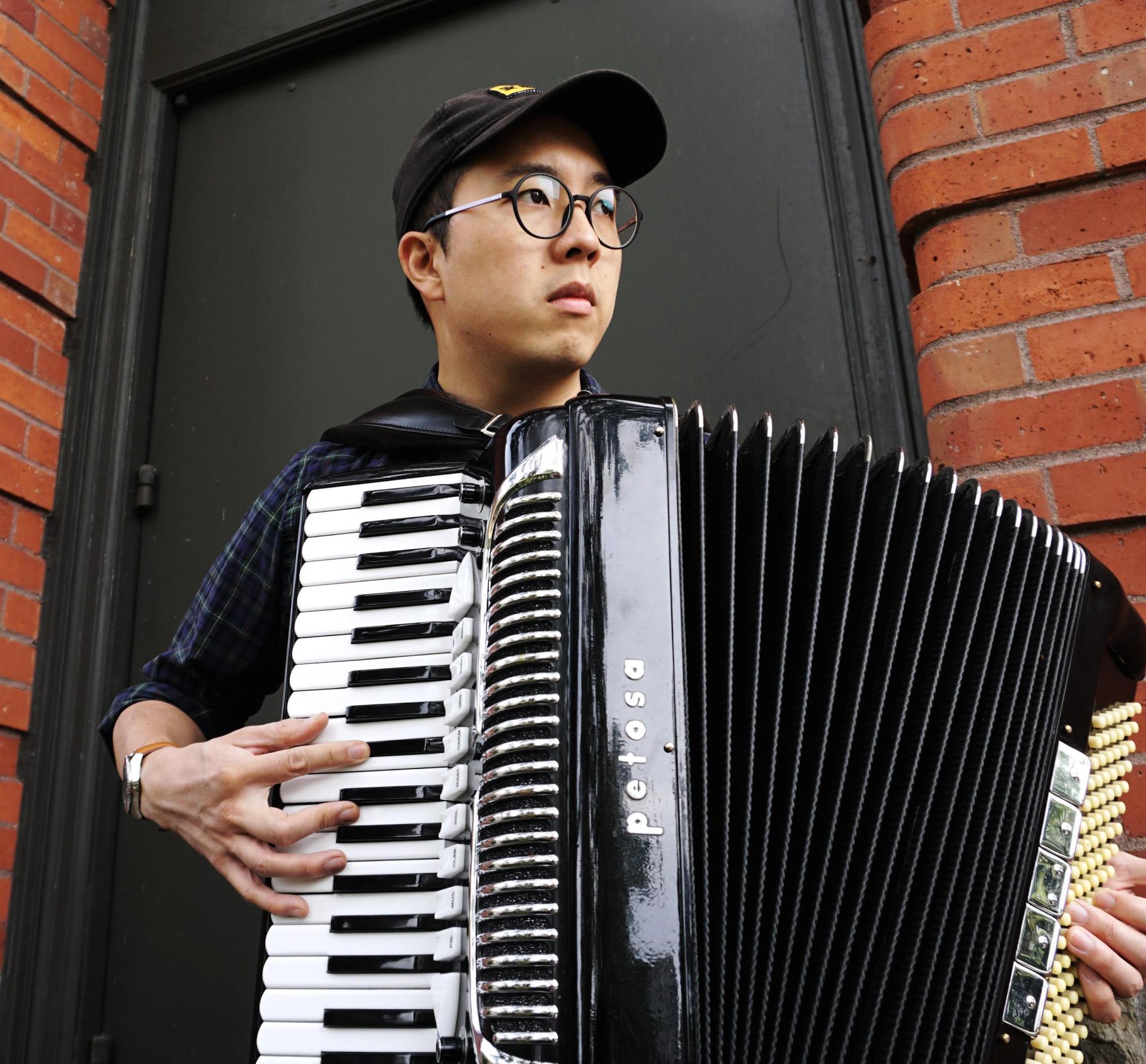 Faculty member Joshua Hou holding an accordion standing in front of a brick wall