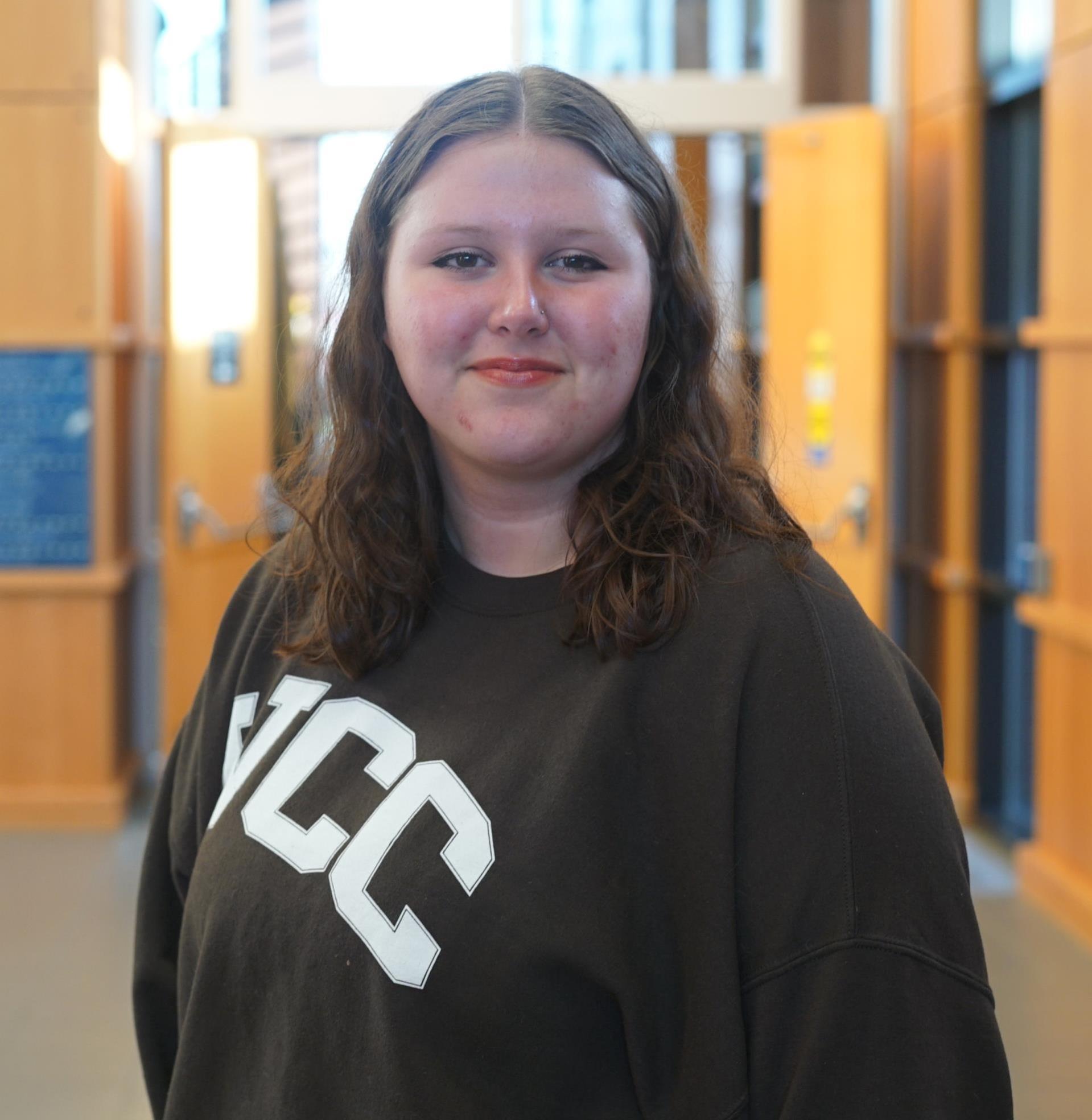 headshot of Kristina standing in front of open doors wearing a brown WCC sweatshirt. She is smiling and has brown hair and brown eyes