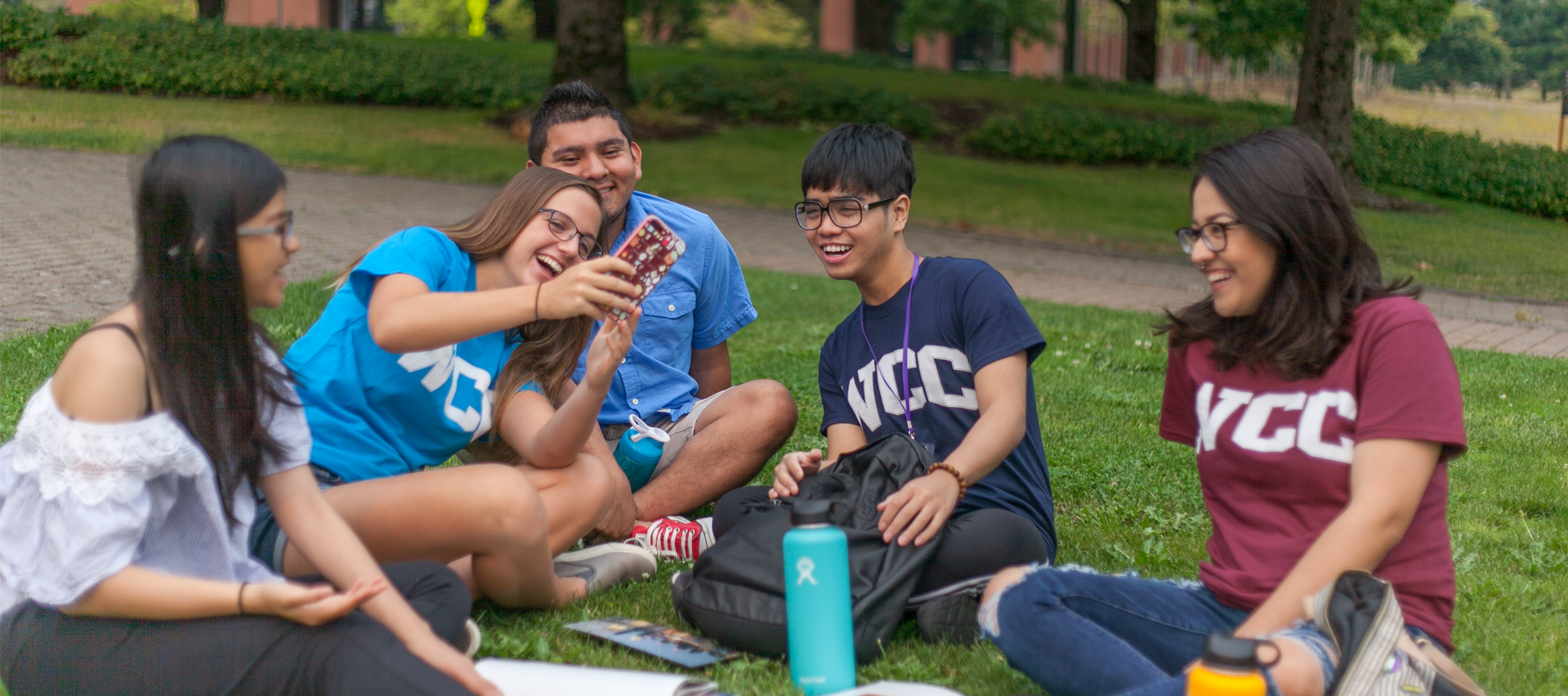 Five WCC students sitting on the lawn in the summertime looking at something on one of the student's phones.